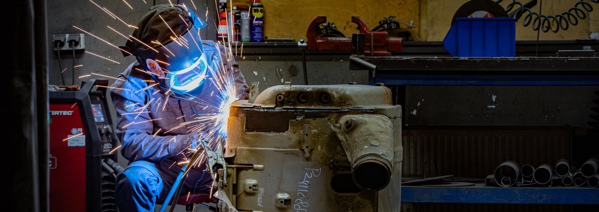 Revisie medewerker aan het werk met het lassen van een uitlaatgassysteem. Hij draagt een lashelm en persoonlijke beschermingsmiddelen